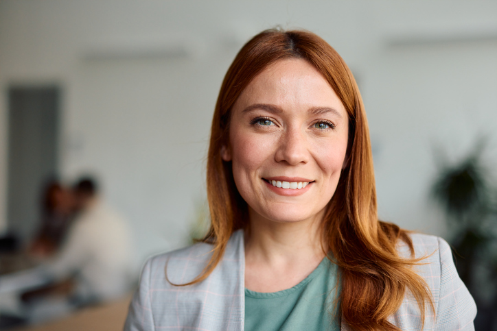 Cheerful redhead businesswoman smiling and engaging with the camera while standing in a blurred office setting, exuding confidence and professionalism in her corporate environment